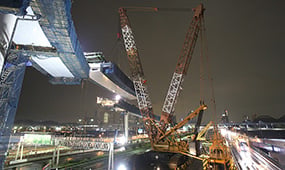夜間の建設現場で、高架橋の巨大な梁を吊り上げて設置する大型クレーン。